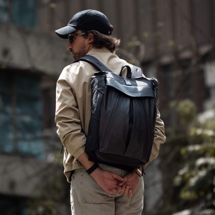 Man wearing a black backpack and cap, standing outdoors with a blurred background